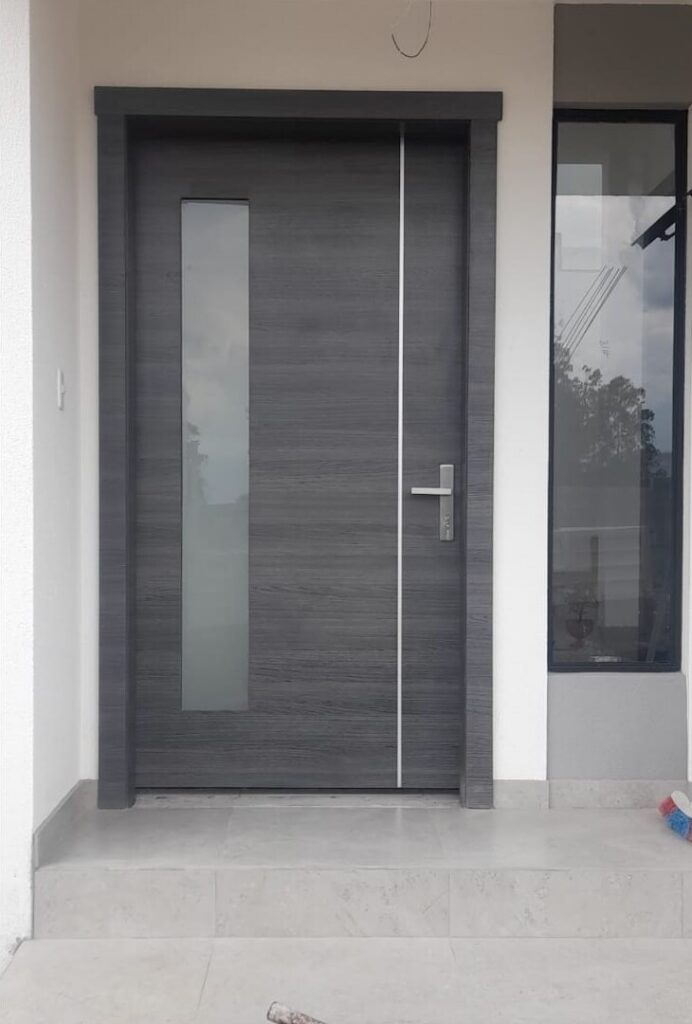 Puerta principal en madera gris con franja de vidrio esmerilado, tirador de acero y ventanal lateral