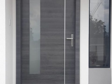 Puerta principal en madera gris con franja de vidrio esmerilado, tirador de acero y ventanal lateral